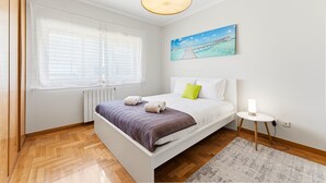 A bedroom with a bed, a bedside table, a lamp, a window with blinds, a radiator, and a painting on the wall. - GuestReady - Charming retreat with balcony in Gaia (Vila Nova de Gaia)