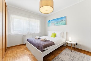Ein Schlafzimmer mit Bett, Nachttisch, Lampe, einem Fenster mit Jalousien, einer Heizung und einem Bild an der Wand.