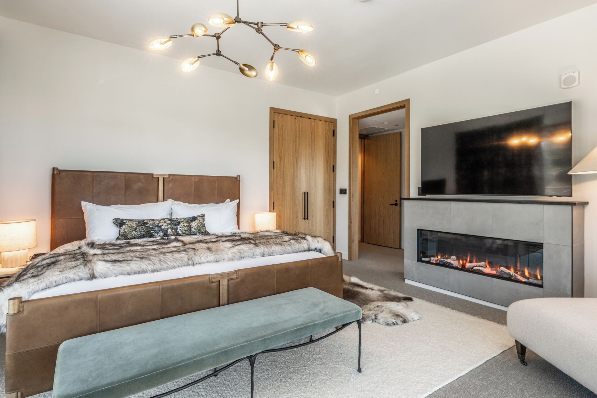 Quarto moderno com uma cama grande, um banco, lareira e televisão na parede.