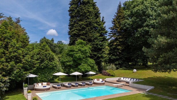 Une piscine entourée de chaises longues et de parasols, elle-même bordée d’une pelouse bien entretenue et de grands arbres.