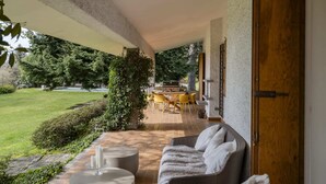 Un espace extérieur aménagé, abrité, avec un canapé, une table et des chaises, un barbecue, et une vue sur une pelouse et des arbres bien entretenus.