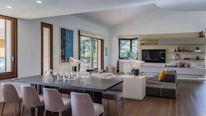 Une cuisine moderne dotée d’un grand îlot central, un coin repas avec une longue table et des chaises, et un espace de vie avec un canapé et un téléviseur.