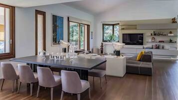 Una cucina moderna con una grande isola, una zona pranzo con un tavolo lungo e sedie, e un soggiorno con divano e televisore.