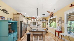 Une cuisine dotée d’un îlot central, de planchers en bois, de meubles blancs, d’un meuble de couleur turquoise et offrant une vue sur l’extérieur grâce à une grande fenêtre.
