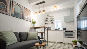 Ein modernes Wohnzimmer mit grauem Sofa, Couchtisch und einem gemusterten Boden. Im Hintergrund ist eine Küche mit weißen Schränken und einem Einbauofen zu sehen.