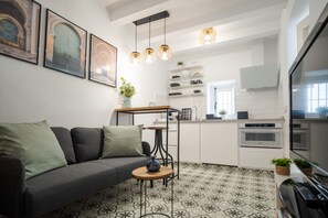 Ein modernes Wohnzimmer mit grauem Sofa, Couchtisch und einem gemusterten Boden. Im Hintergrund ist eine Küche mit weißen Schränken und einem Einbauofen zu sehen.