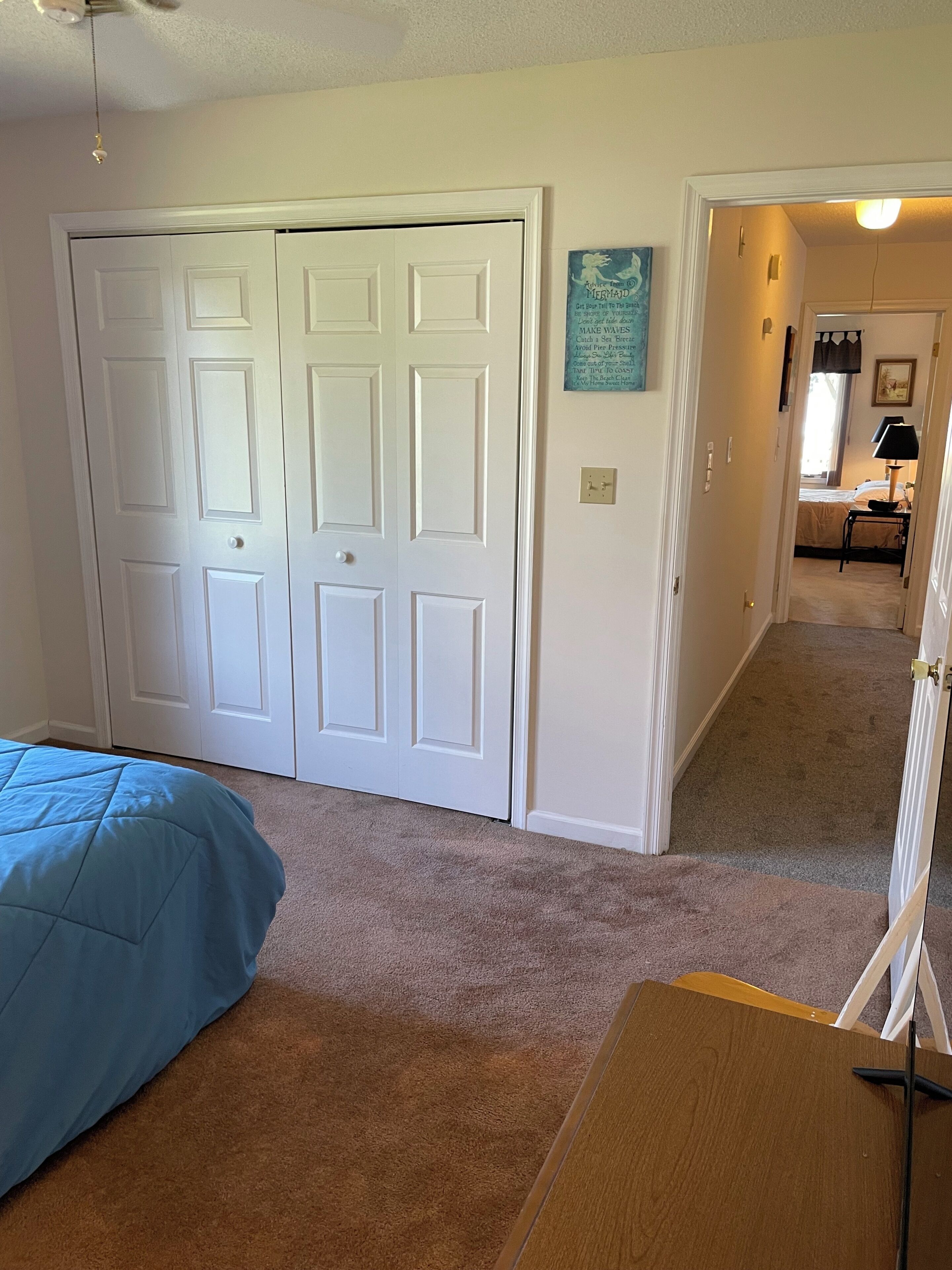 A bedroom with a bed, a closet, a hallway leading to another room, and a wall with a framed quote.