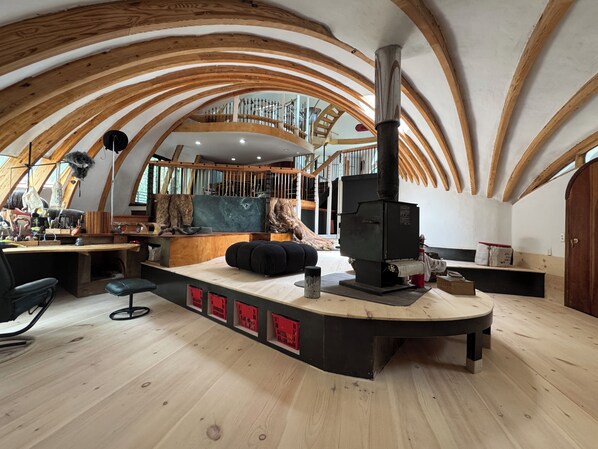 A rustic interior with a wooden ceiling, a central fireplace, a workbench, and a seating area.