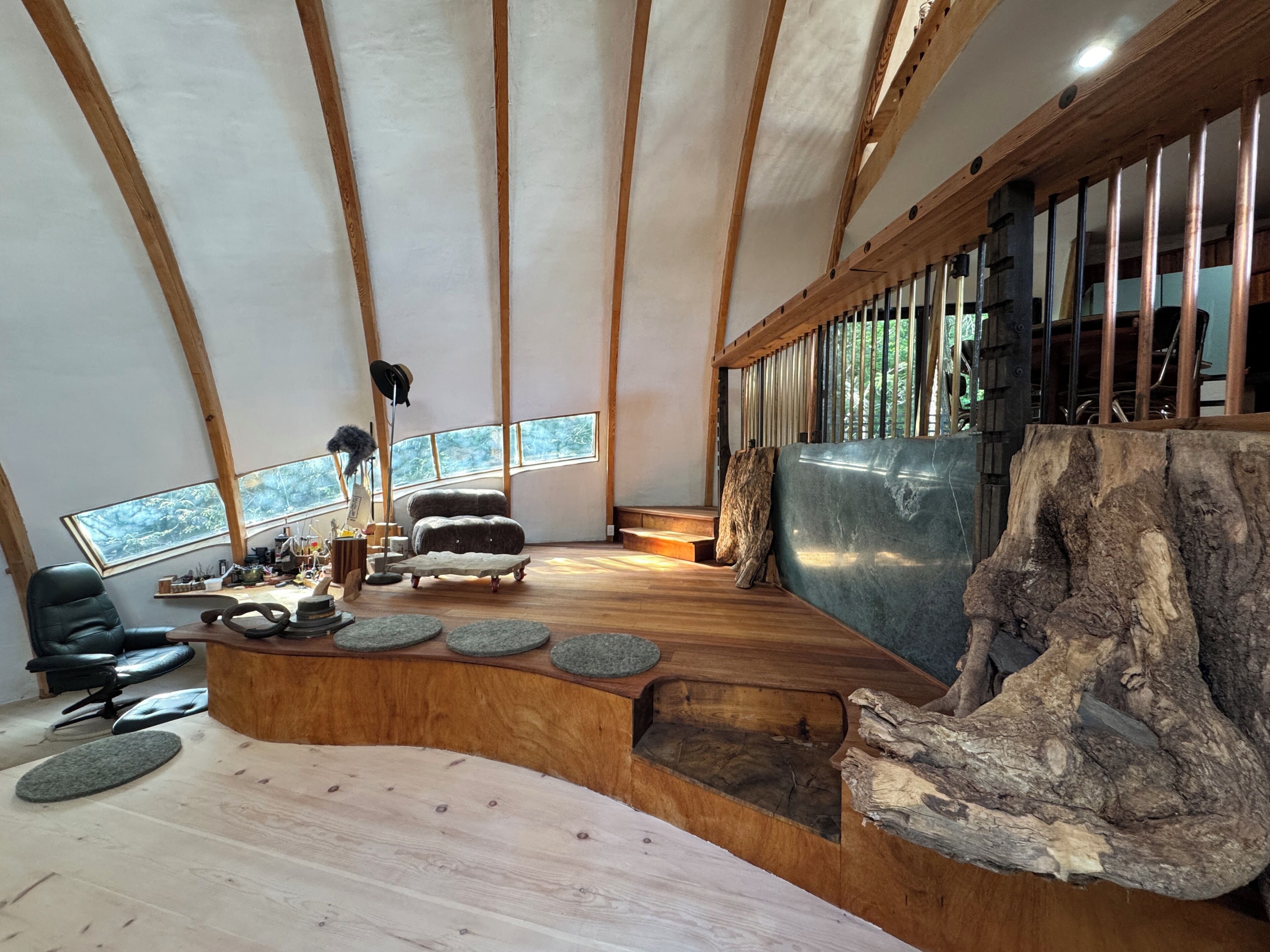Una habitación con un banco de madera curvo, un grueso tronco de árbol y una ventana que da a vistas de árboles.