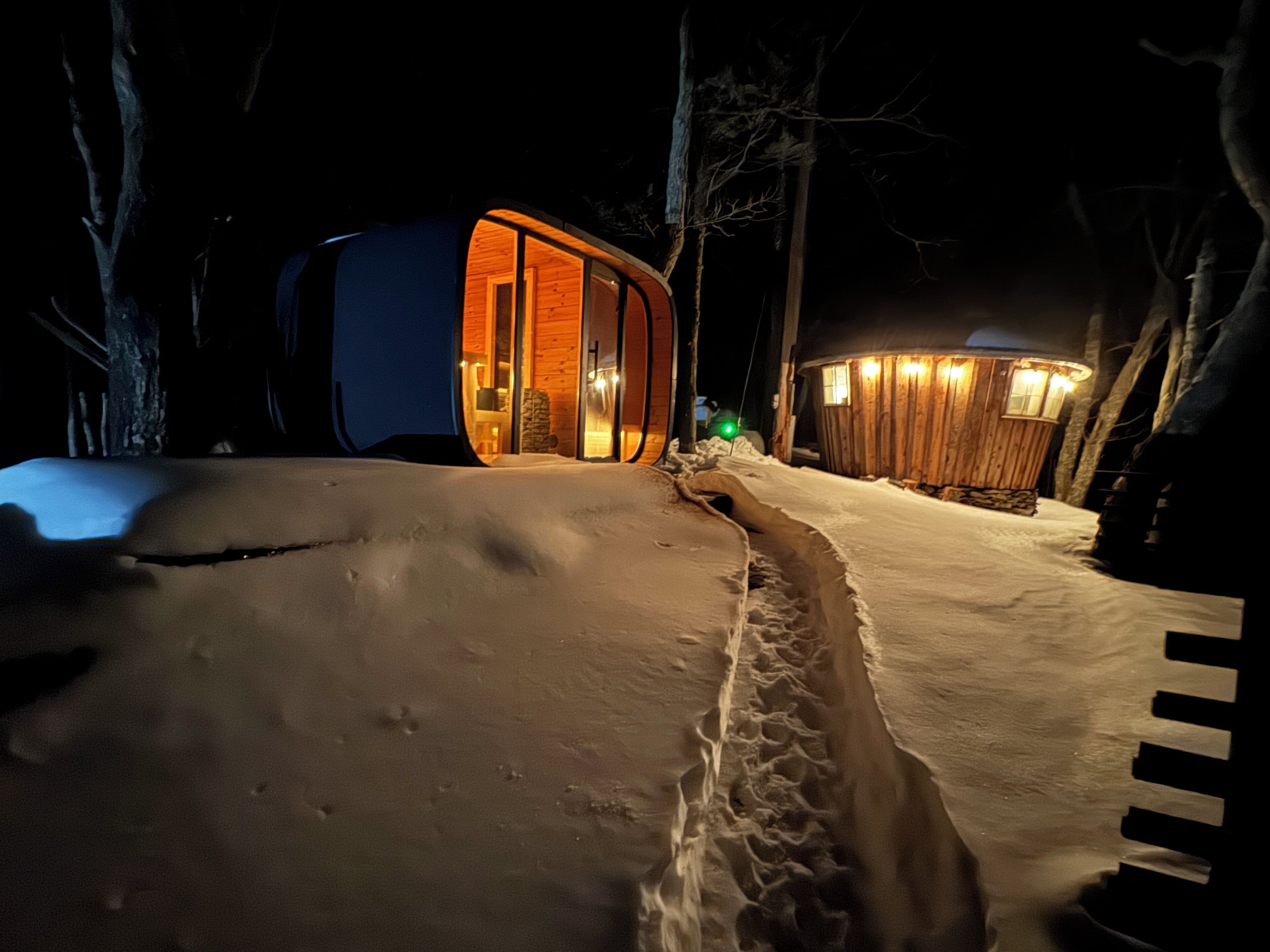 Ein verschneiter Weg führt zu einer Holzhütte mit Glastür und beleuchtetem Innenraum.