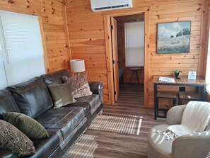 A wooden cabin interior with a leather sofa, a small table, a chair, and a painting on the wall. - Cozy 2-Bedroom Cabin Near Two Kings Casino & Local Attractions (Grover)