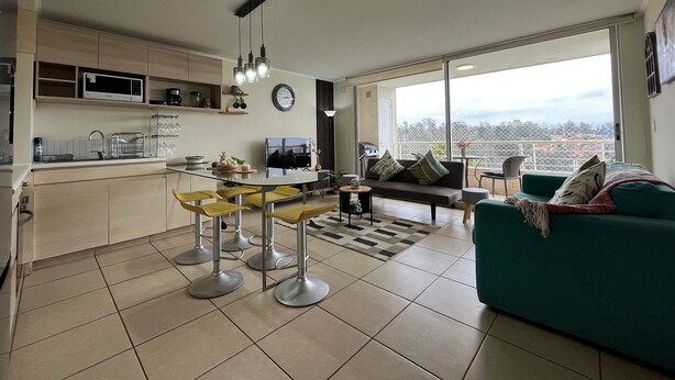 A modern living space with a kitchen, dining area, and a large window offering a city view. - Oceana Suites Laguna Bahía (Algarrobo)