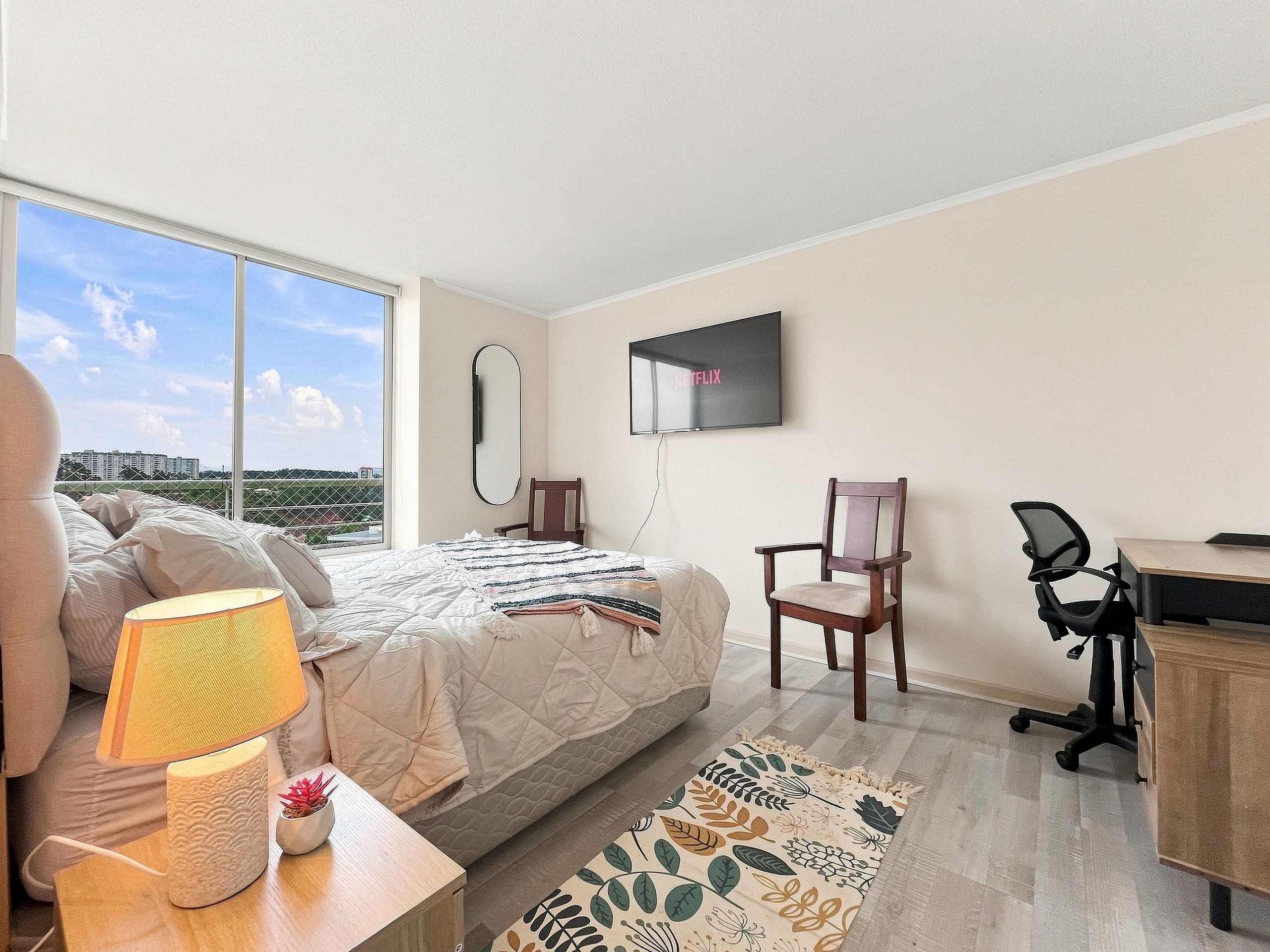Un dormitorio con cama, escritorio, silla, televisor y vistas de la ciudad.