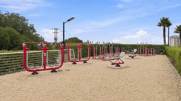 An outdoor fitness area with exercise equipment, a sandy surface, and a metal fence.