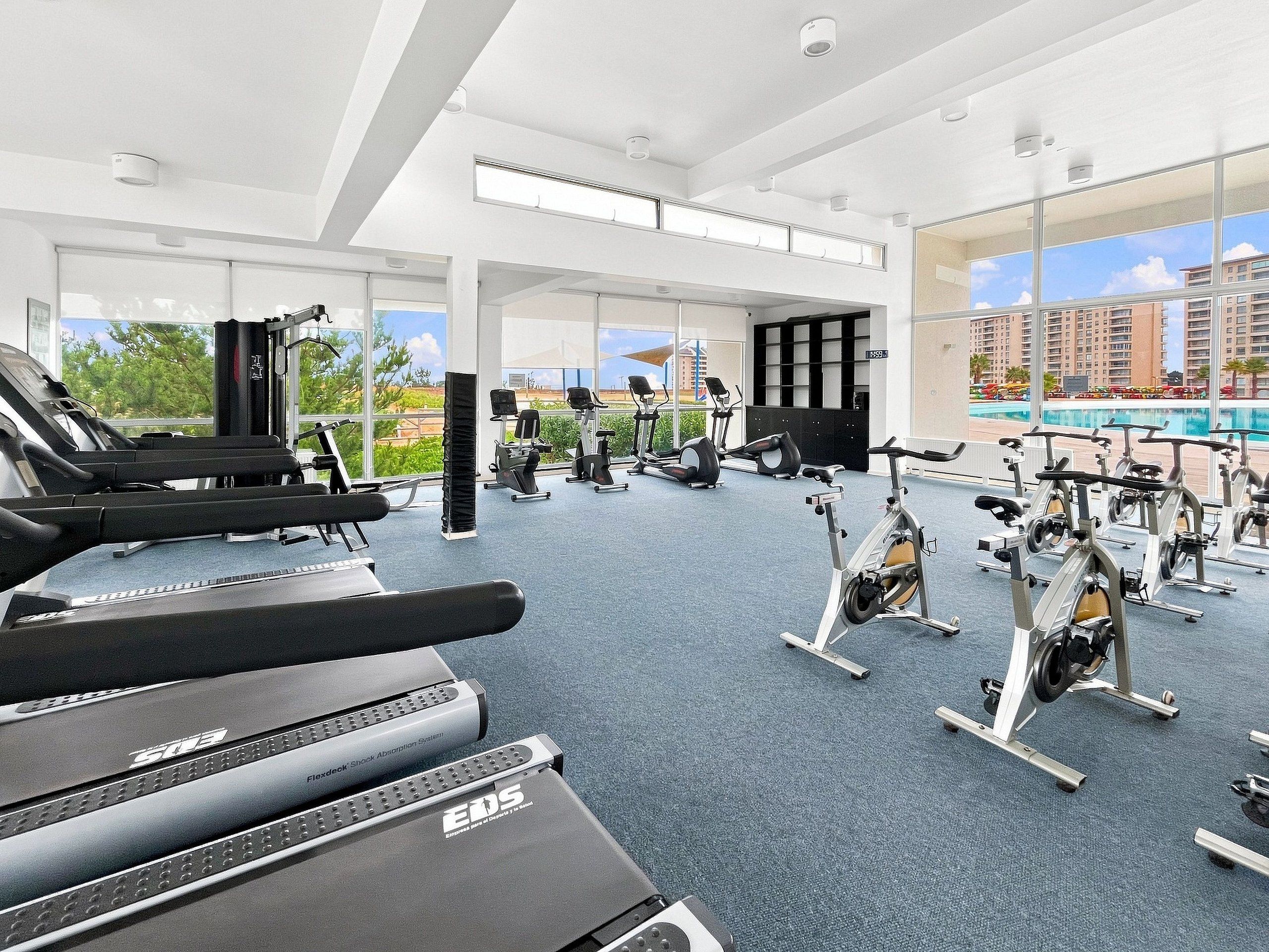 Ein Fitnessstudio mit Cardiogeräten, Kraftstationen und einem großen Fenster mit Blick auf Gebäude und Grünflächen.