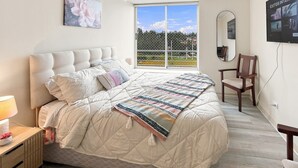 A bedroom with a large bed, a bedside table with a lamp, a chair, a window with a view, and a wall-mounted TV. - Oceana Suites Laguna Bahía (Algarrobo)