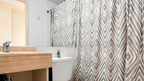 A bathroom with a wooden vanity, a toilet, and a shower curtain with a geometric pattern. - Oceana Suites Laguna Bahía (Algarrobo)