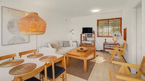 Un salon comprenant un canapé, une table basse, des fauteuils, un luminaire suspendu, une bibliothèque et une fenêtre avec des stores.