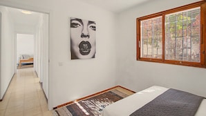 Une chambre à coucher avec un lit, une grande fenêtre avec des stores, une porte donnant sur une autre pièce et une œuvre d’art accrochée au mur.