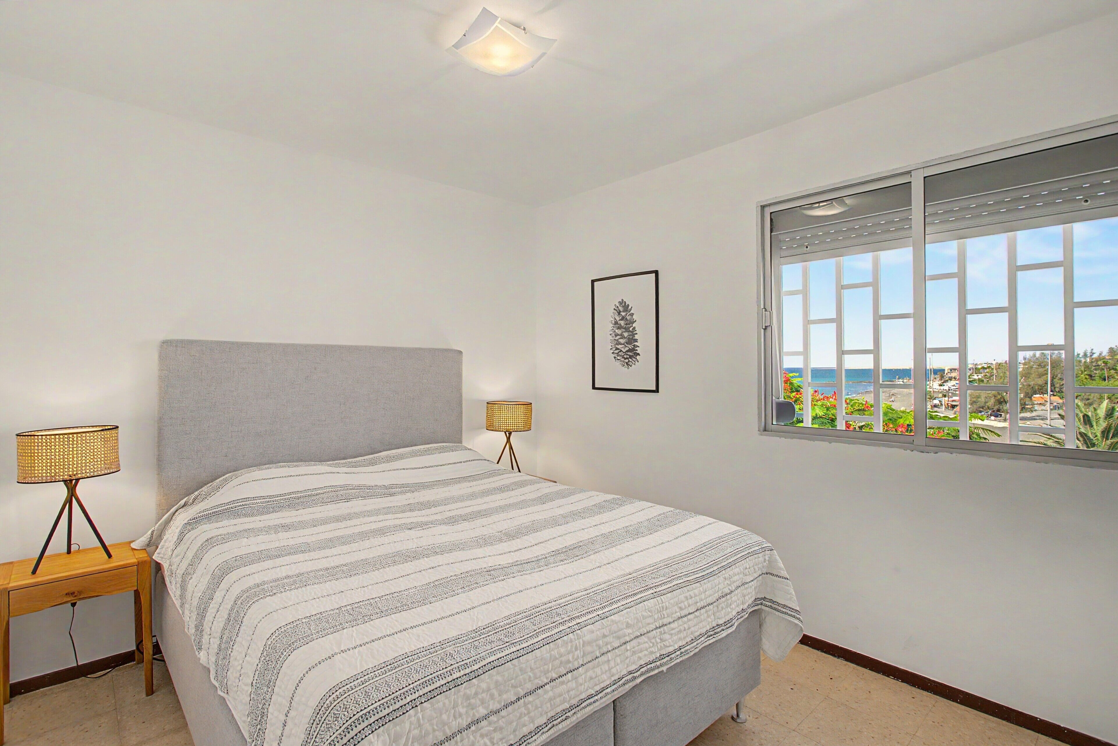 Ein Schlafzimmer mit einem Bett, einer Nachttischlampe, einem gerahmten Bild und einem Fenster mit Blick.