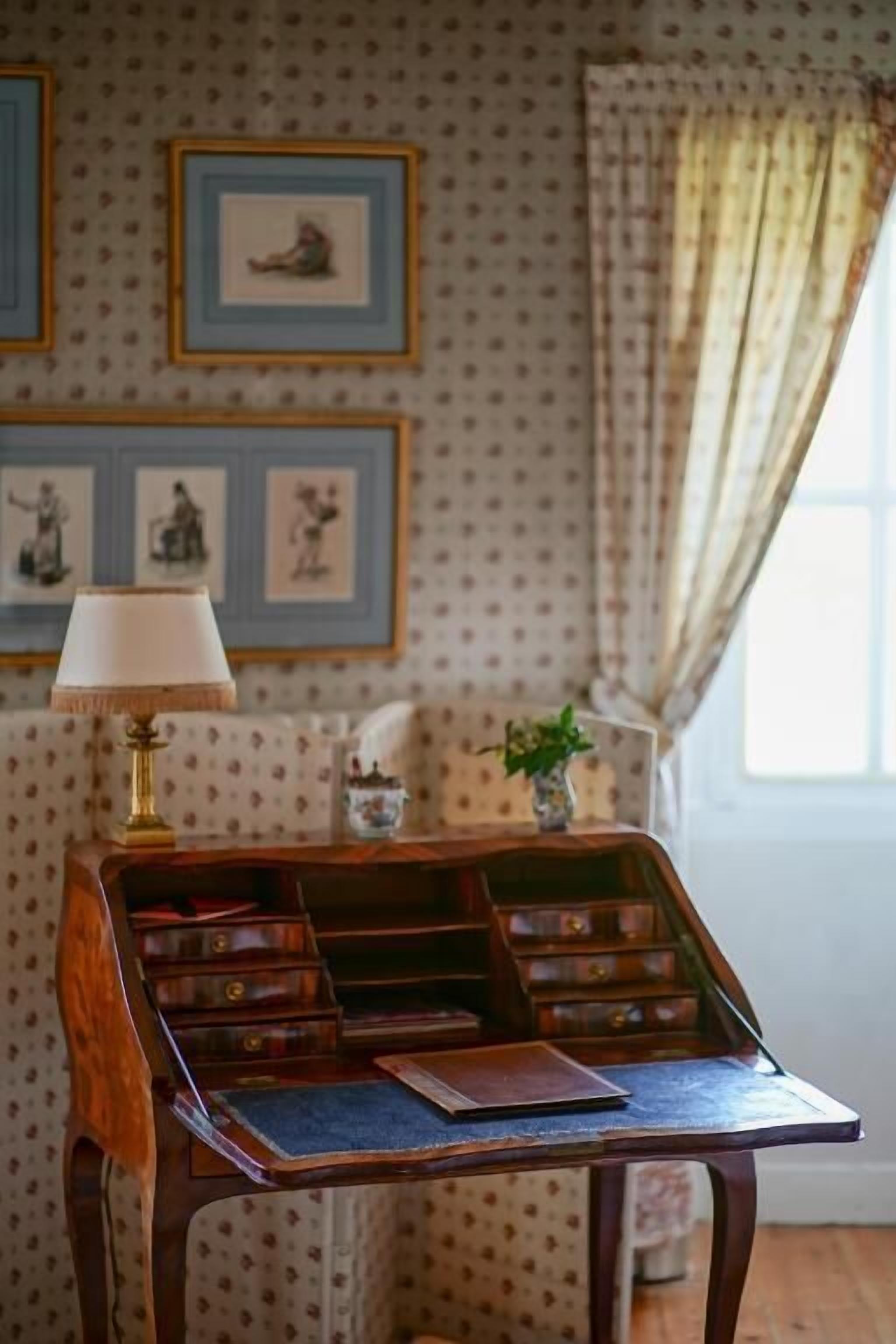 Een houten bureau met lades en een lamp, een lijst met een foto aan de muur, en een raam met gordijnen.