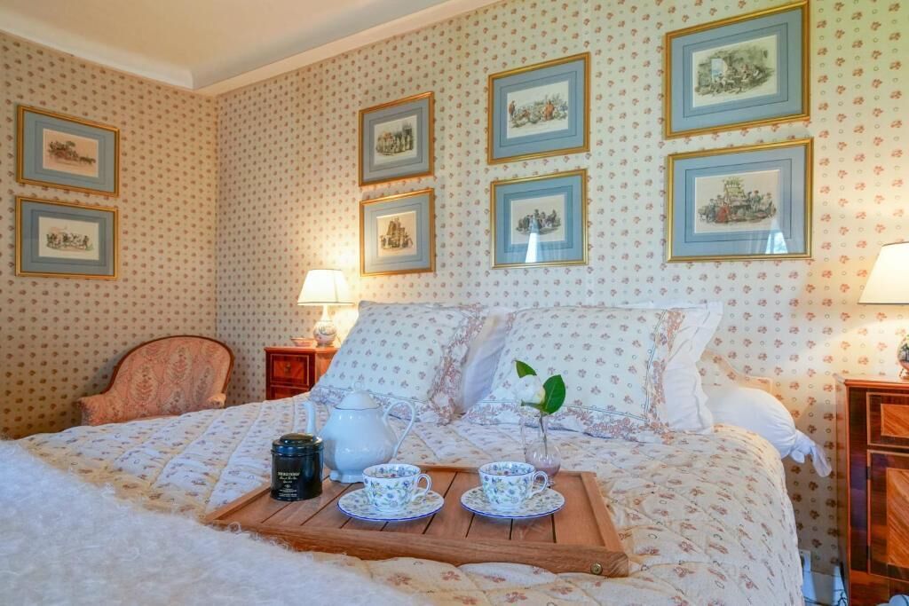 A bedroom with a bed, a bedside table with a lamp, a wooden tray with teacups and a teapot, and framed pictures on the wall.