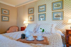 Un dormitorio con una cama, una mesita de noche con una lámpara, una bandeja de madera con tazas de té y una tetera, y cuadros en la pared.