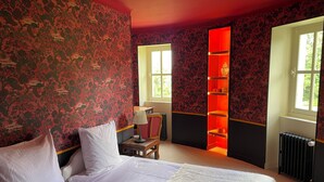Habitación de hotel con cama, mesita de noche, lámpara, sillón, ventana y estante iluminado con luz roja.
