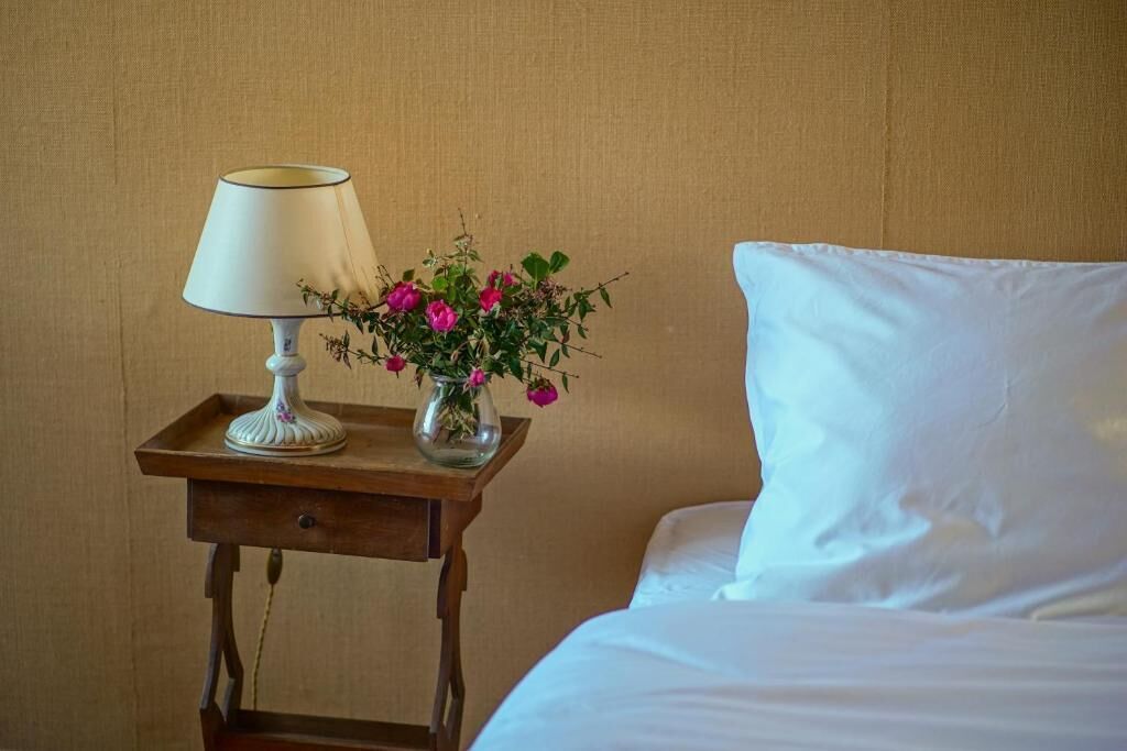 A bed with white sheets, a bedside table with a lamp, and a vase with flowers.