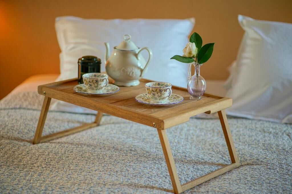 A wooden tray with a teapot, cups, and a vase with a flower on a bed.