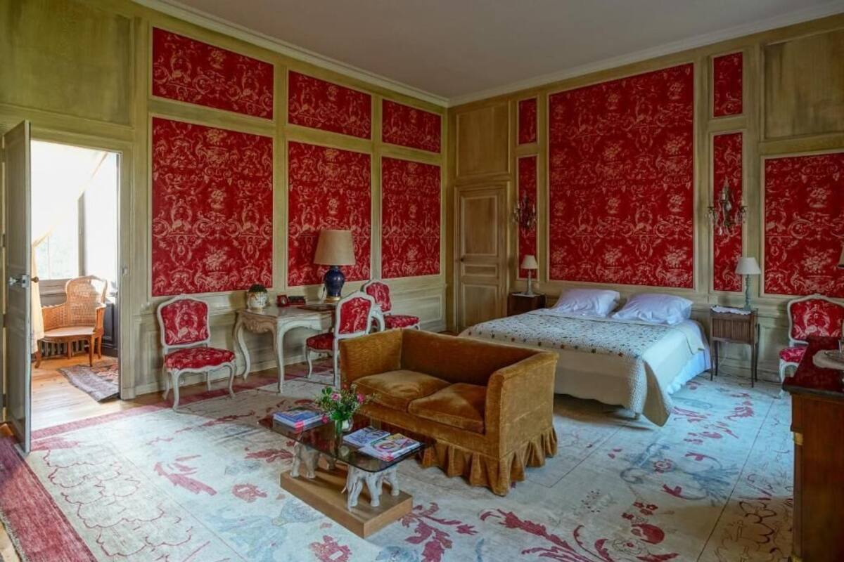 Una habitación con una cama, un sofá, una silla, una mesita con un jarrón y una puerta que da a otra habitación.