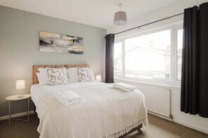 Habitación de hotel con cama, mesita de noche, lámpara, ventana con cortinas y un cuadro en la pared.
