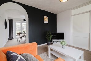Ein Wohnzimmer mit schwarzer Wand, einem weißen Sofa, einem weißen Couchtisch, einem Fernseher und einem durch einen Bogen sichtbaren Essbereich.
