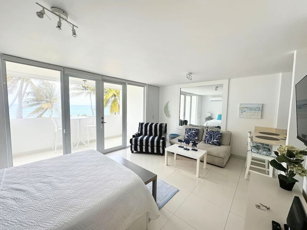 Una habitación de hotel moderna con una cama grande, un sofá, una mesa de centro, un televisor y un balcón con vistas a palmeras.