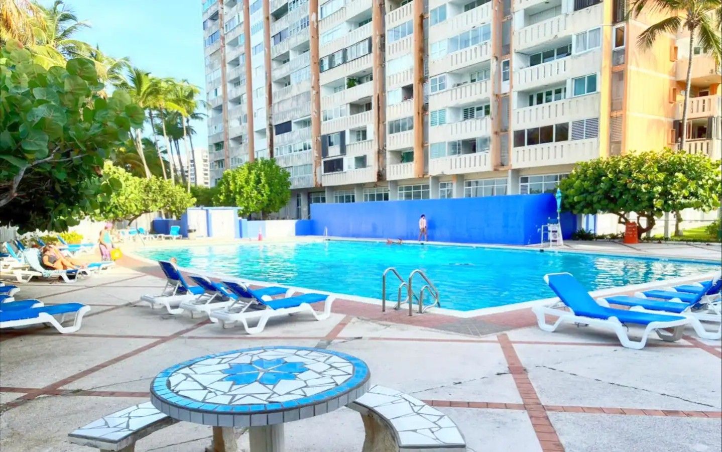 Una piscina con lettini, un solarium piastrellato e un grattacielo sullo sfondo.