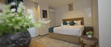 A hotel room with a bed, a small table with a plant, a nightstand, and a wall-mounted air conditioning unit.