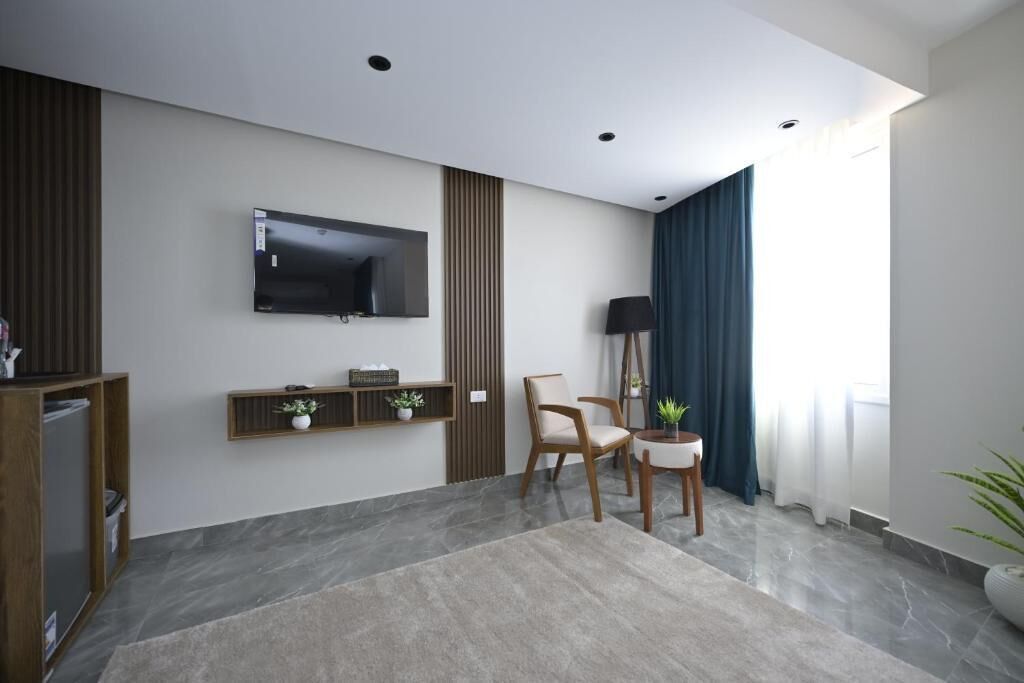 Una sala de estar moderna con una televisión de pantalla plana montada en la pared, un mueble de televisión de madera con objetos decorativos, un par de sillones, una lámpara de pie y un gran ventanal con cortinas.