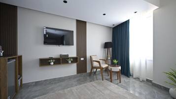Una sala de estar moderna con una televisión de pantalla plana montada en la pared, un mueble de televisión de madera con objetos decorativos, un par de sillones, una lámpara de pie y un gran ventanal con cortinas.