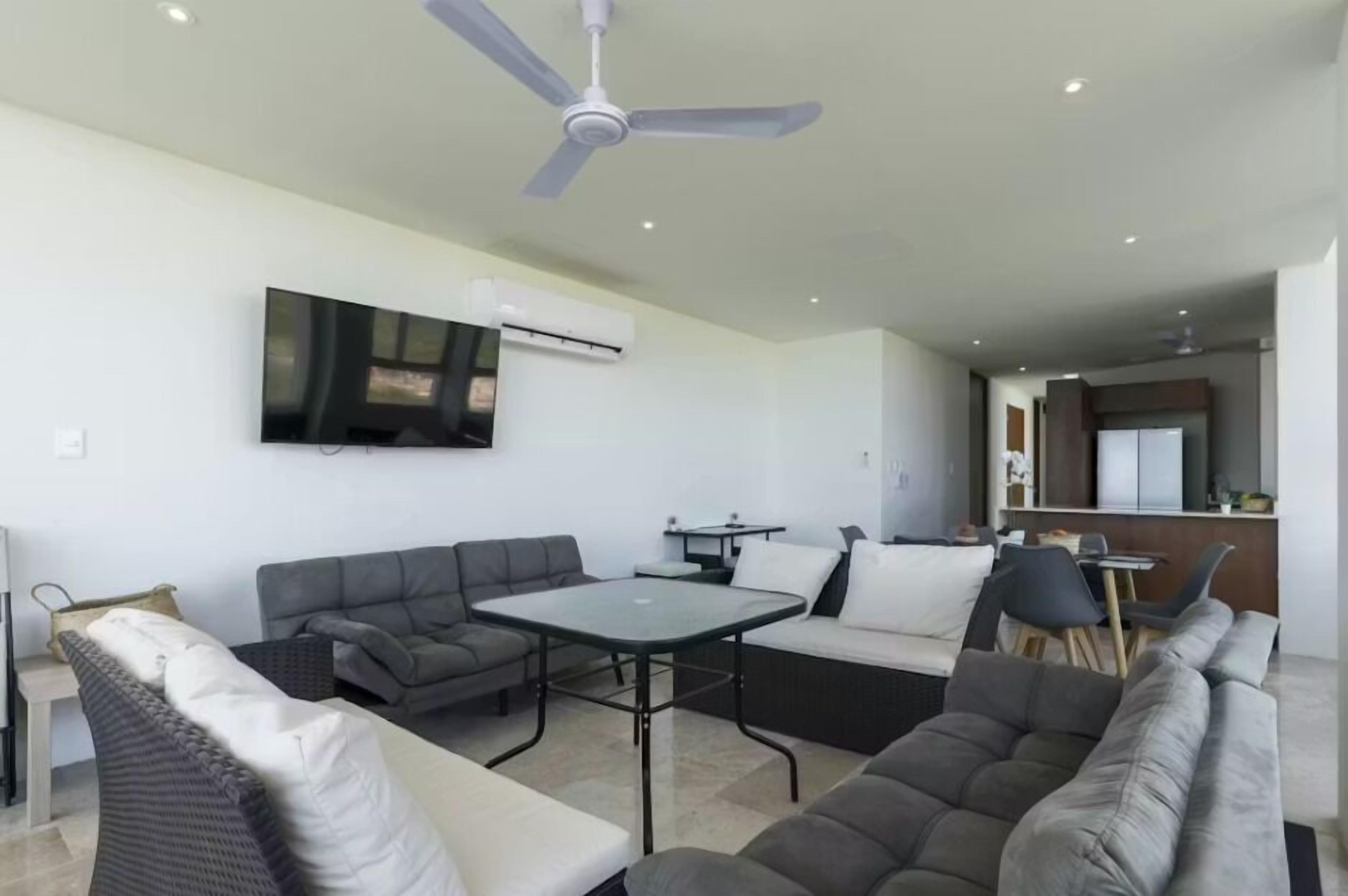 Un salon moderne doté d’un téléviseur à écran plat fixé au mur, d’un ventilateur de plafond et d’un coin repas avec une table et des chaises.