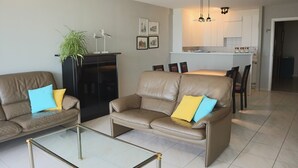 Ein Wohnzimmer mit einem Glastisch, zwei Sofas mit Kissen, einer schwarzen Kommode und einer Küchenzeile im Hintergrund.