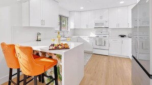 A modern kitchen with white cabinets, a central island with seating, and a dining area with orange chairs. - 4BR Oasis Near MIA - Jacuzzi - Games - BBQ (Miami)
