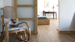 A wooden rocking chair with a colorful cushion, a wooden table with a lamp, and a blue sofa in a room with wooden flooring. - Gîtes de la Soulondre: Vacation Home with Mountain View, Shared Pool, and Wi-Fi (Lodève)