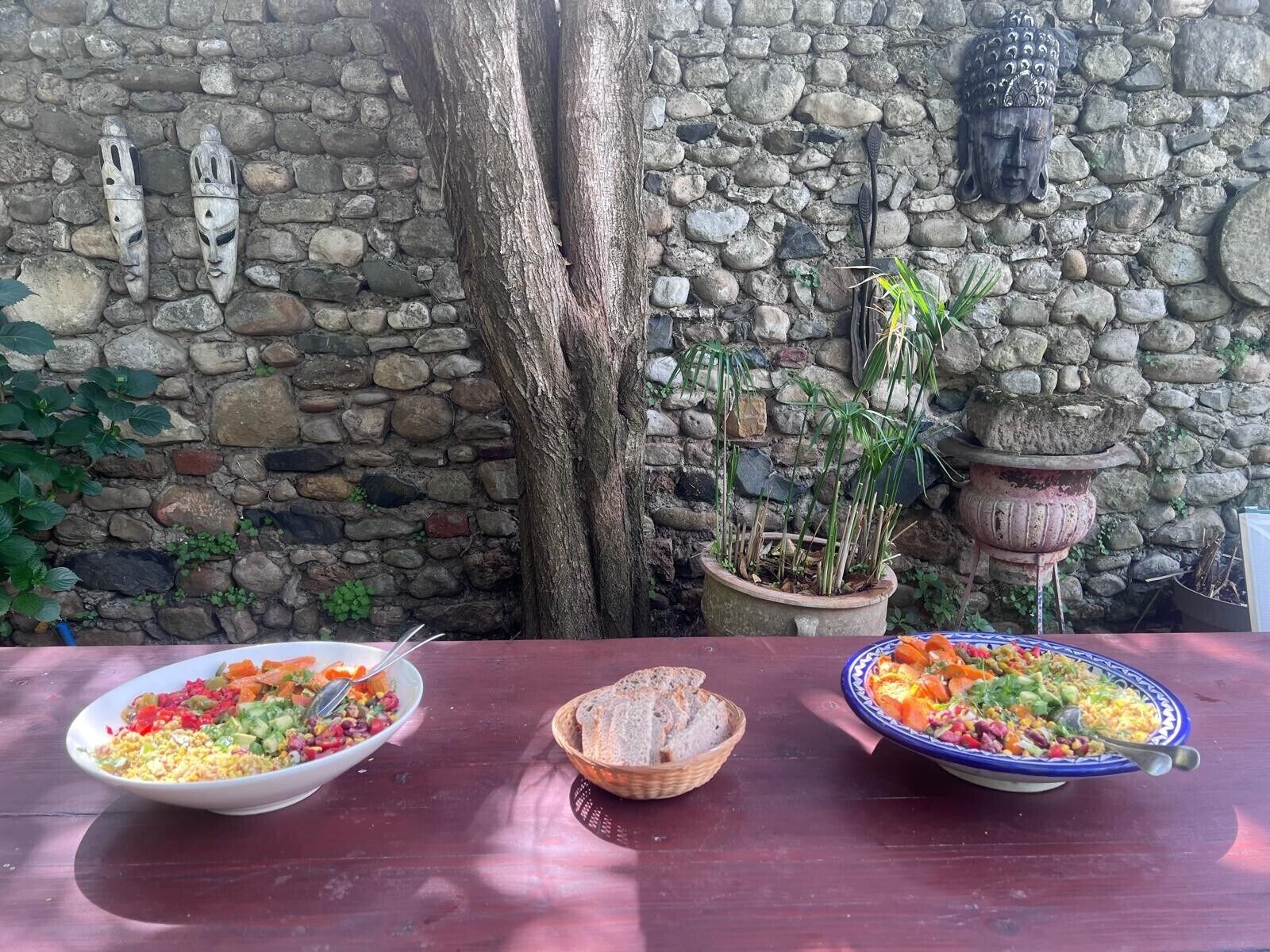 A stone wall with a Buddha head sculpture, a tree trunk, and a table with three bowls of food.