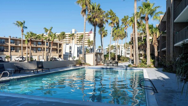 A swimming pool with lounge chairs, surrounded by palm trees and a multi-story building with balconies. - 2BR Half Block to Beach, Pool Hot Tub Patio SPI (South Padre Island)