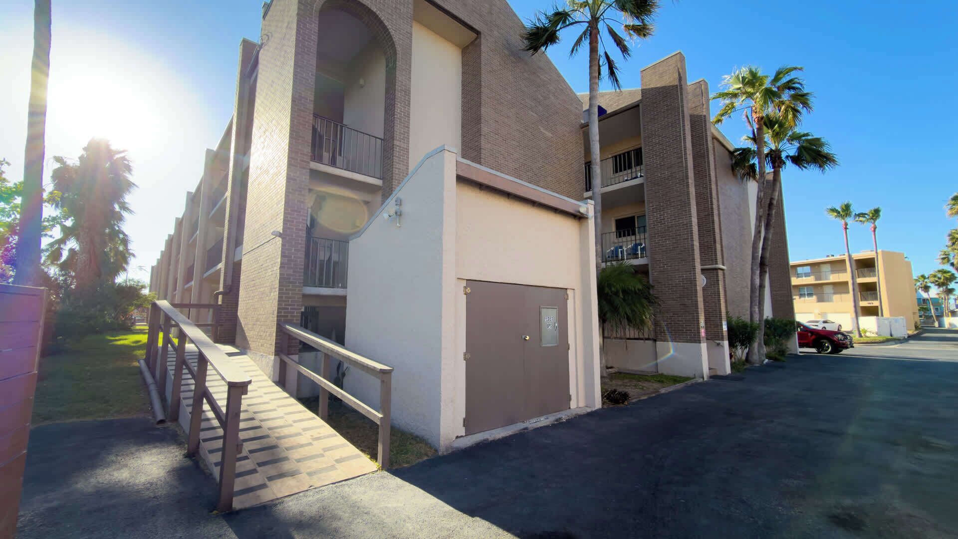Un edificio de varios pisos con una escalera central, balcones y un estacionamiento con palmeras.