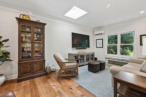 Ein Wohnzimmer mit einem Holzschrank, einem Korbstuhl, einem Ledersitzhocker und einem Fernseher auf einem Standfuß.