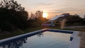 En swimmingpool med en solnedgång i bakgrunden, omgiven av träd och en parkerad skåpbil.