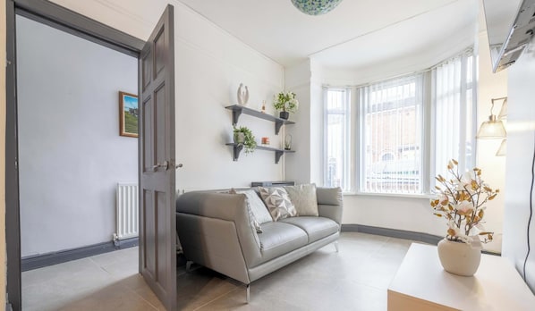 Ein modernes Wohnzimmer mit einem grauen Sofa, einem Heizkörper, einem Fenster mit Jalousien und einem Blumentopf auf einem Tisch.
