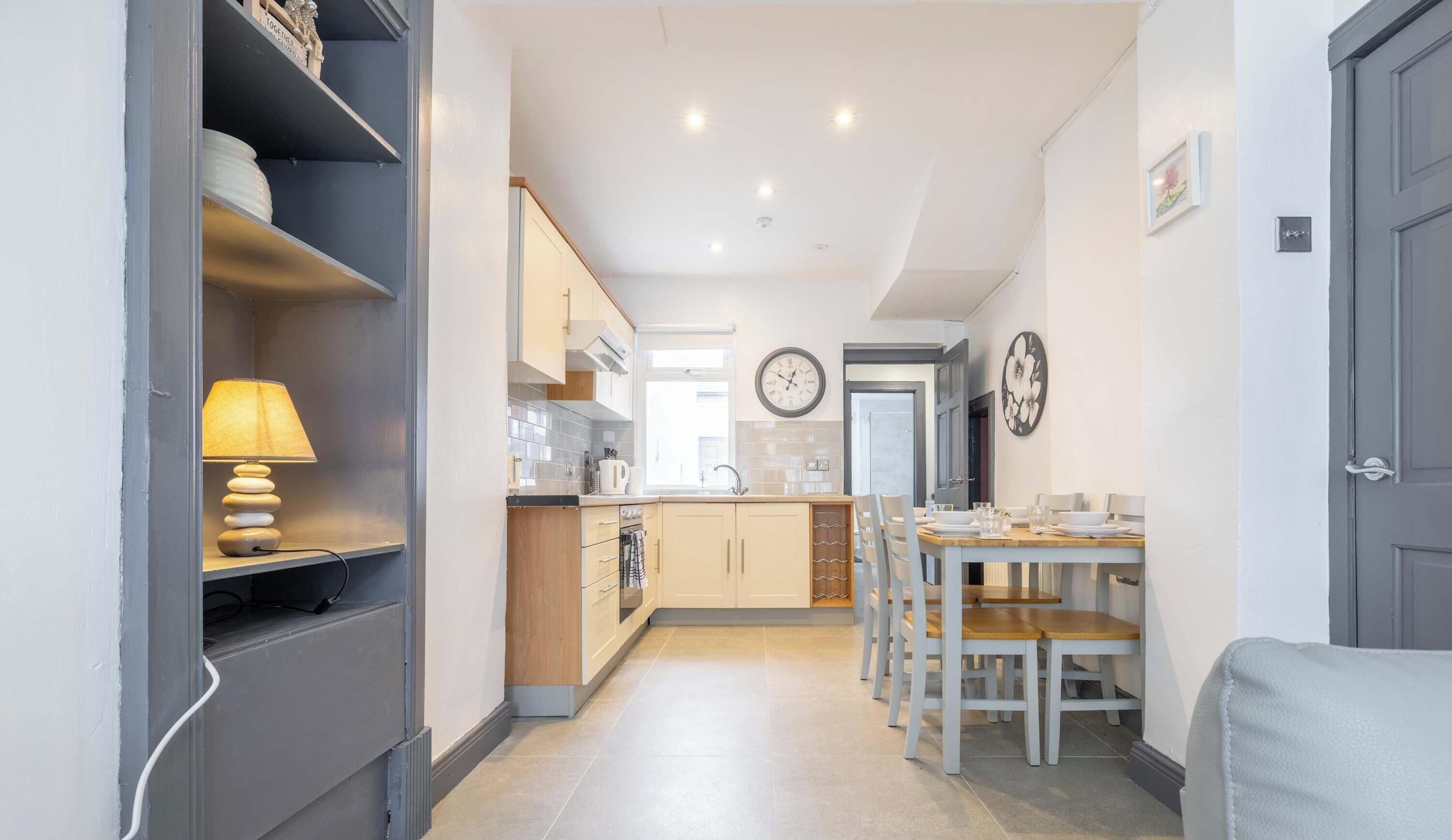 A modern kitchen with a dining area, a clock on the wall, and a sofa in the foreground.