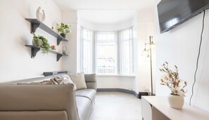 Ein modernes Wohnzimmer mit einer Couch, einem an der Wand befestigten Fernseher und einem Fenster mit Blick auf Gebäude.